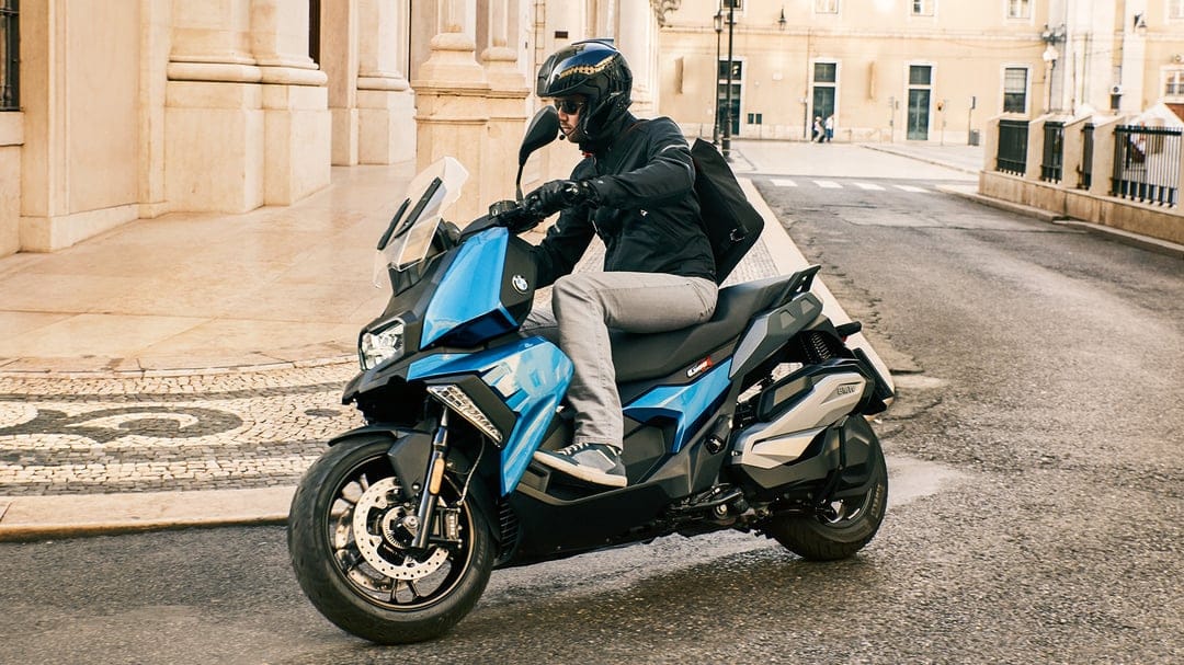 Blue BMW scooter being ridden by a man in a black helmet on a sunlit historic urban street with stone buildings.