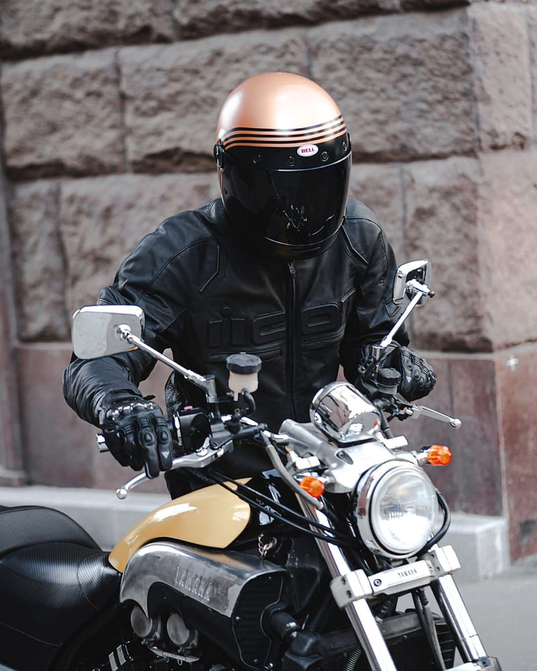 Motorcyclist in a copper helmet and black leather jacket sits on a vintage yellow Yamaha bike with chrome parts.