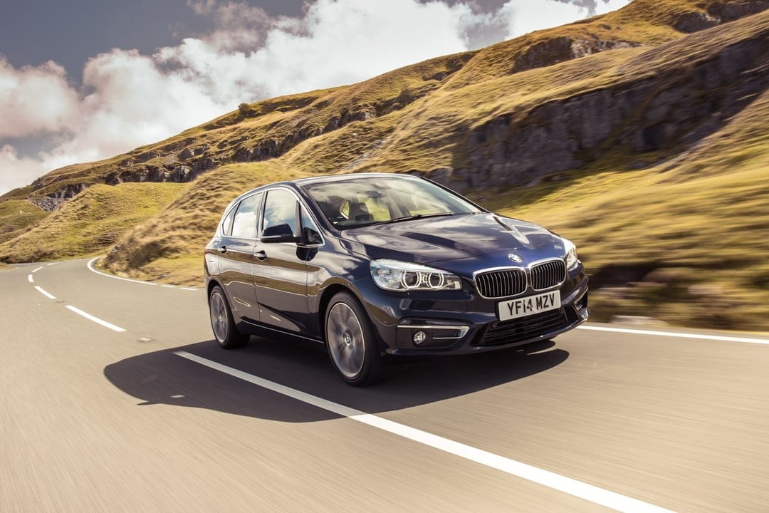 Dark blue luxury car driving on a winding mountain road with motion blur and sunlit grassy hills in the background.