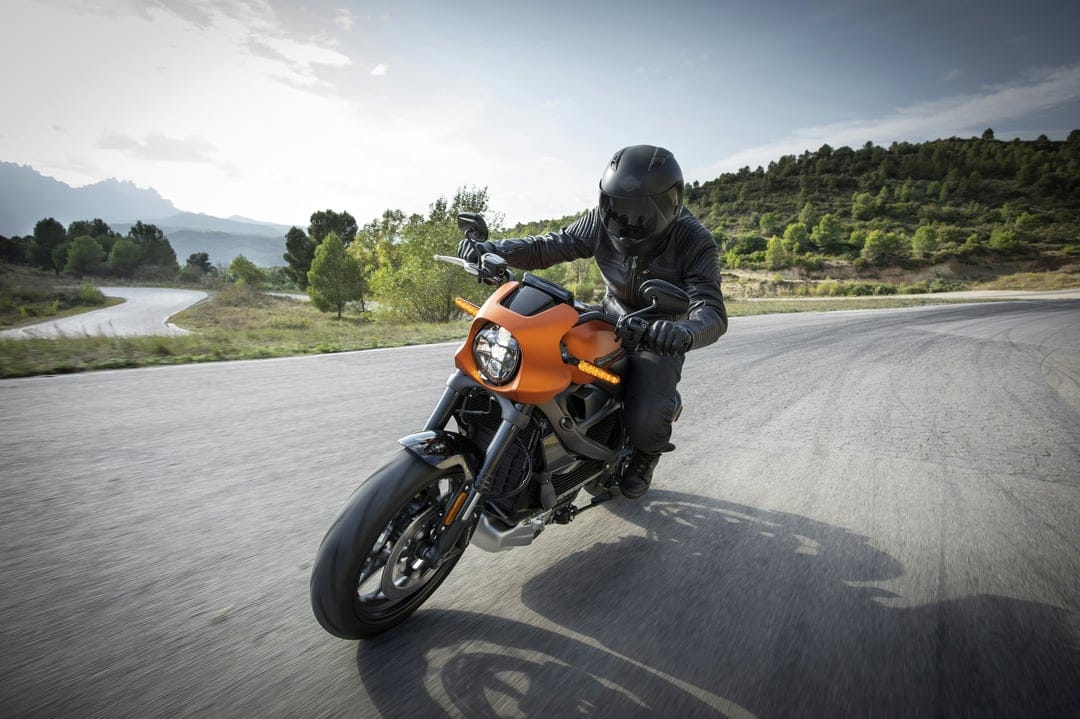 Motorcyclist on an orange sportbike leans into a turn on a paved road with green hills under a blue sky.