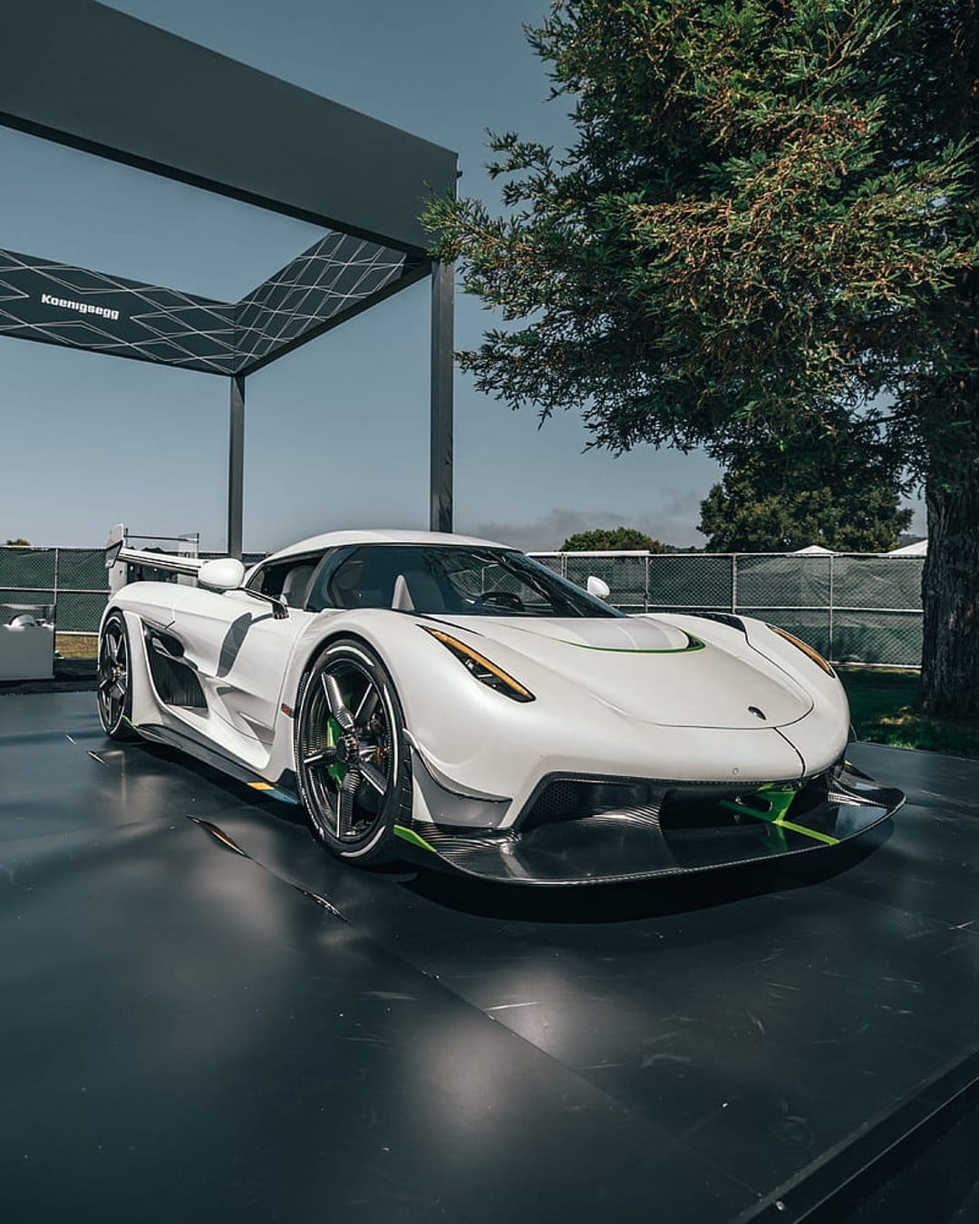 Koenigsegg Jesko hypercar in white with green accents parked on a dark platform near a modern glass building.