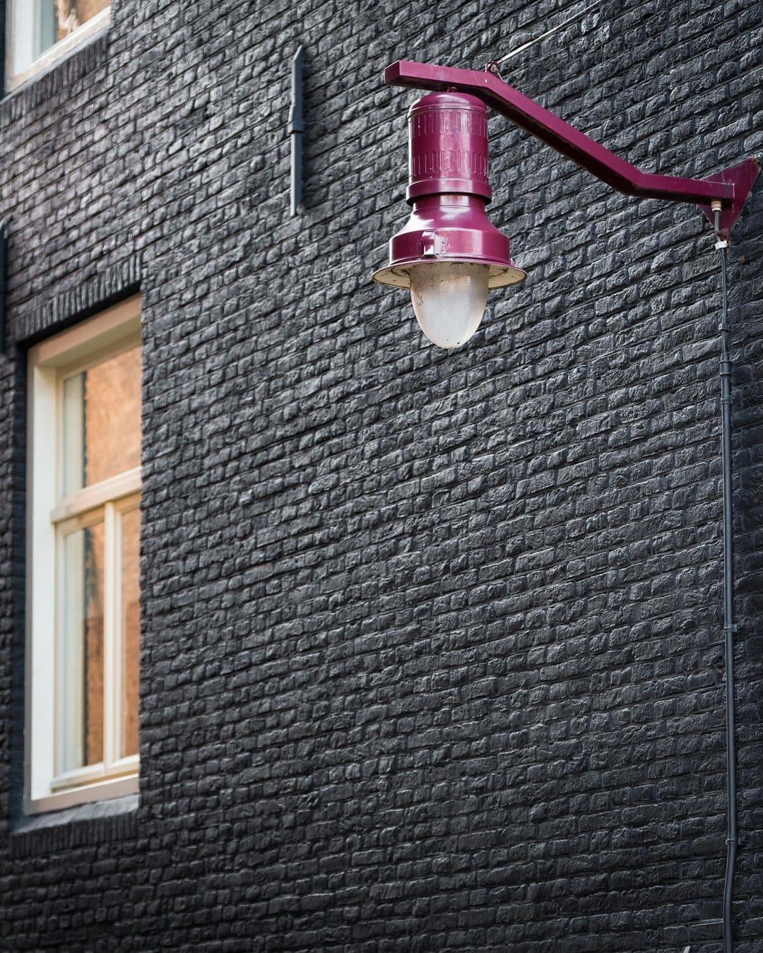 Purple vintage street lamp mounted on a textured black brick wall beside a cream window frame in vertical view.