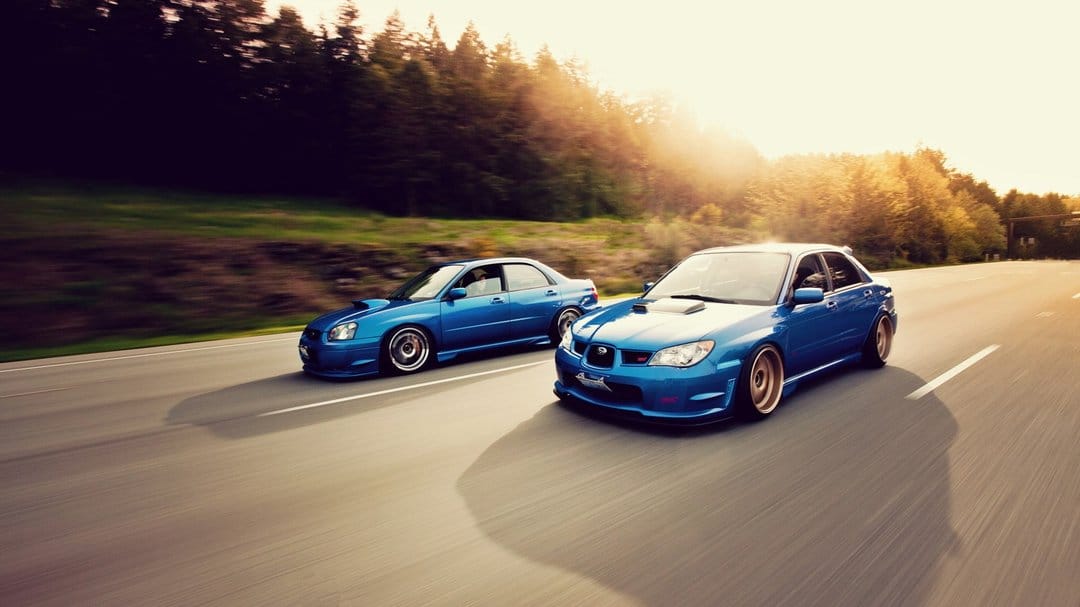 Two blue Subaru WRX STI cars driving on a sunlit highway with motion blur and forest background at sunset.