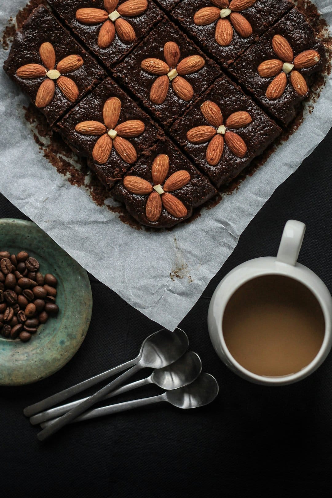 Chocolate brownies topped with almond petals sit beside a steaming coffee mug and roasted beans on parchment.