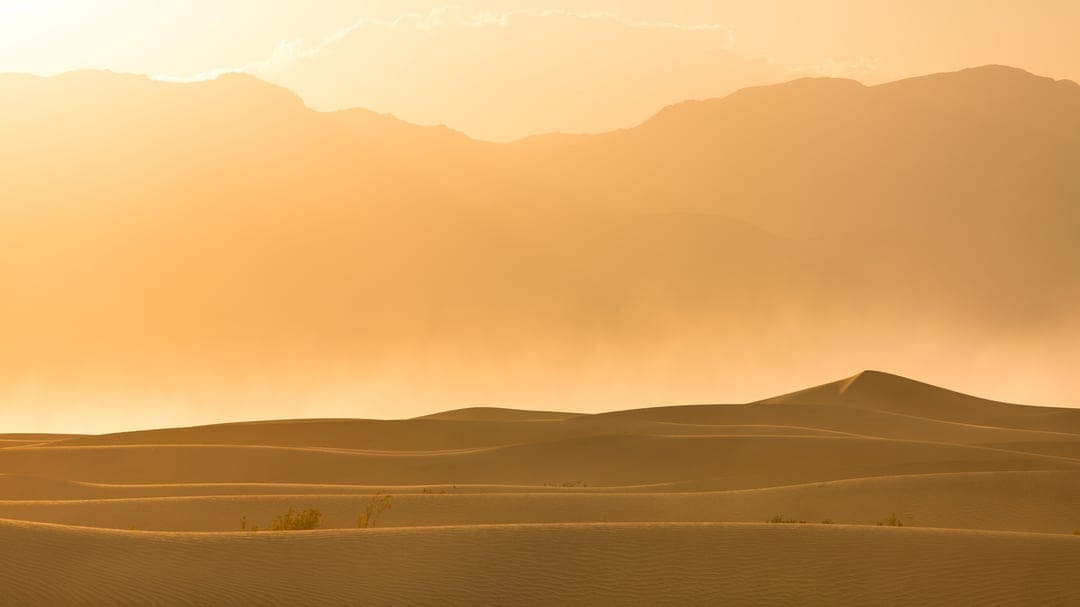 Undulating sand dunes and silhouetted mountain peaks glow under a hazy golden hour sun with floating dust motes.