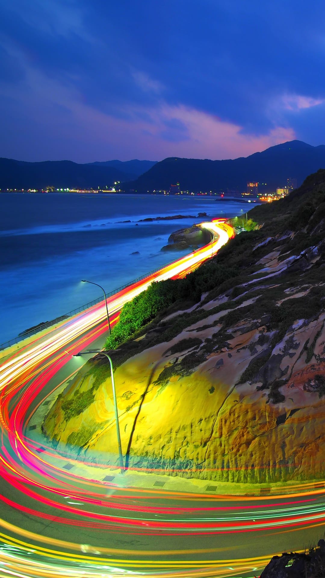 Glowing red and white light streaks curve along a coastal highway beside a rocky cliff and ocean at dusk.