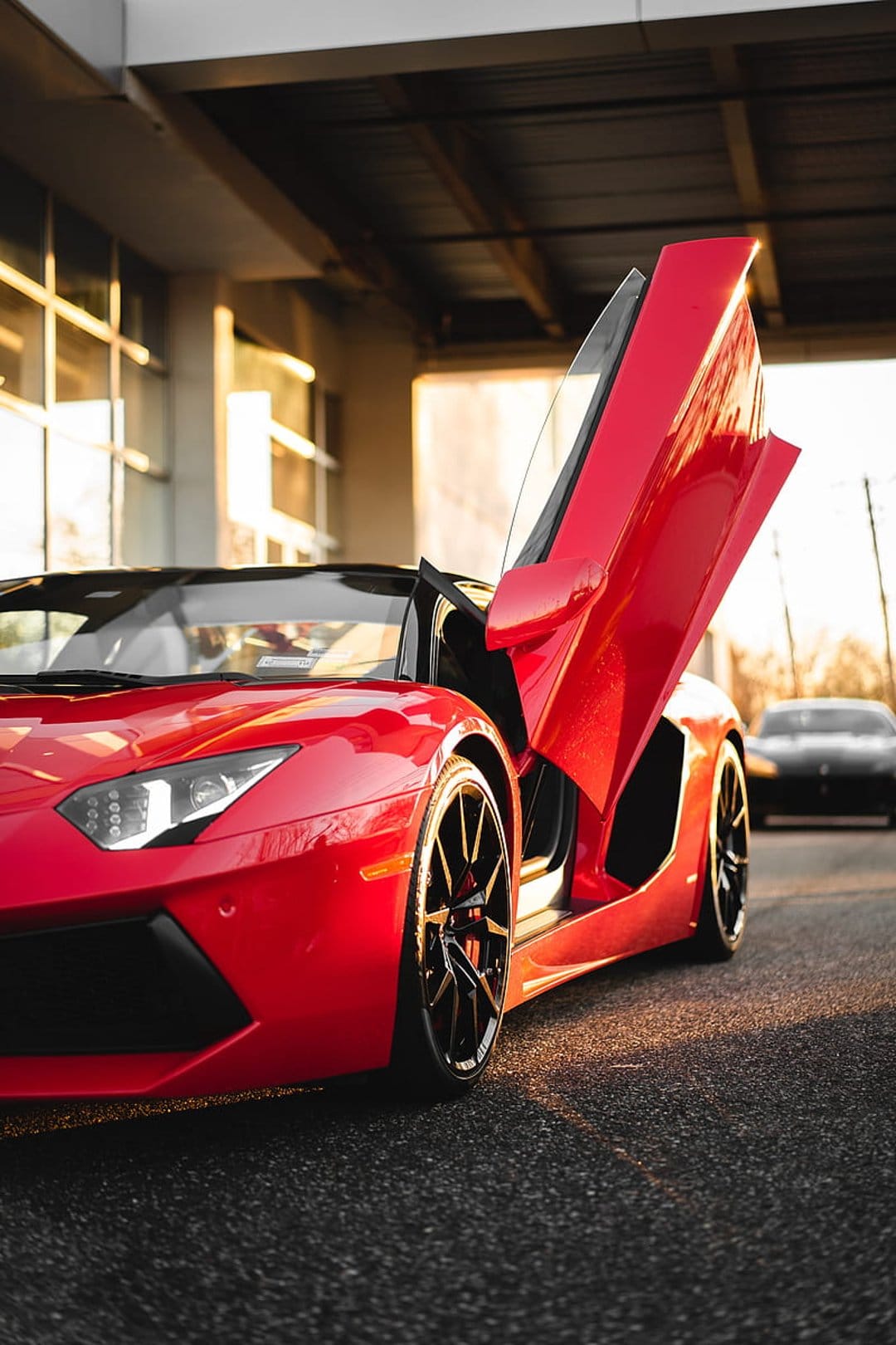 Red Lamborghini Aventador with open scissor doors and black rims parked on grey asphalt in bright sunlight.