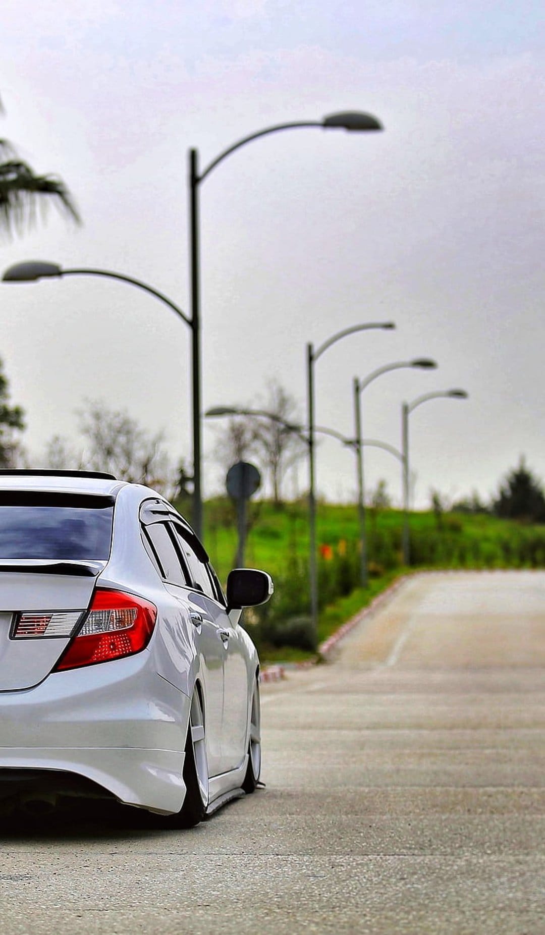 White sedan with a lowered suspension parked on a paved road beside tall streetlights and lush green trees.