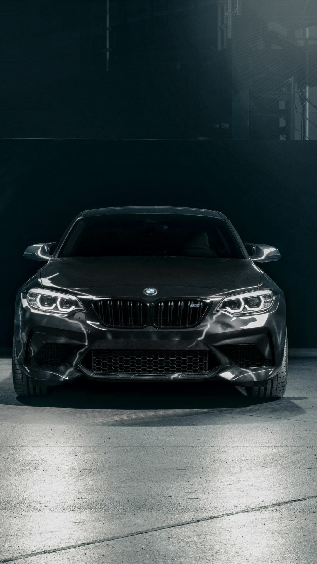 Front view of a dark grey BMW M2 with glowing white LED headlights in a dimly lit studio setting.