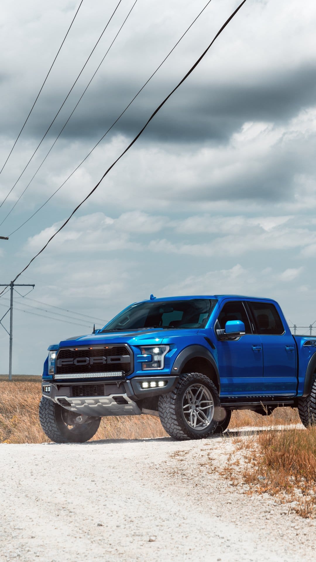 Blue Ford Raptor truck parked on a white gravel path under a dramatic cloudy sky with power lines crossing above.
