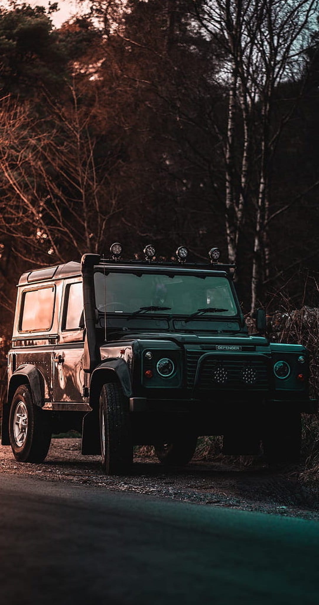 Black Land Rover Defender with off-road gear parked on a dirt forest trail during a vibrant sunset glow.