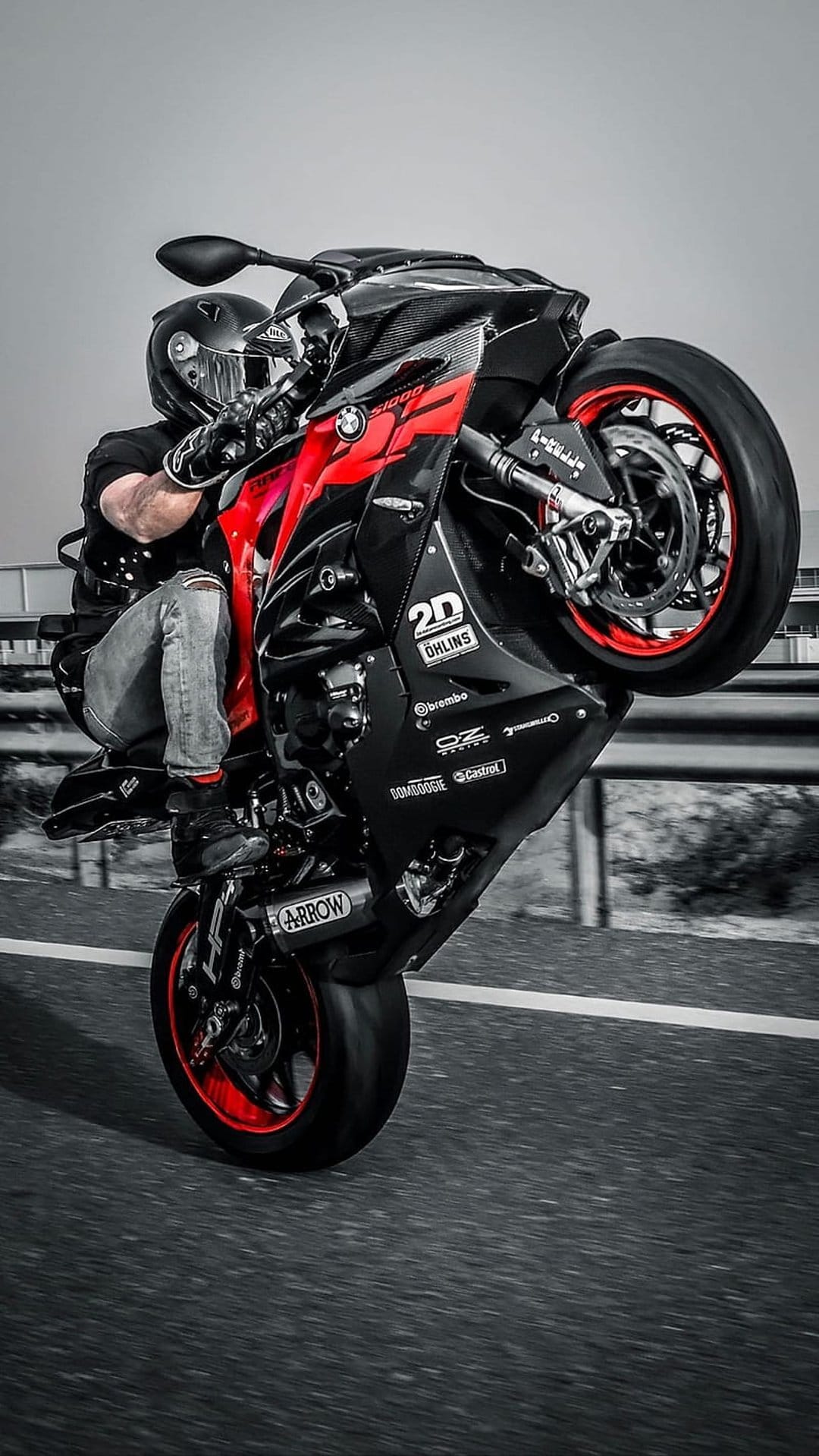Motorcyclist in racing gear performs a high wheelie on a red and black sportbike across a gray asphalt road surface.