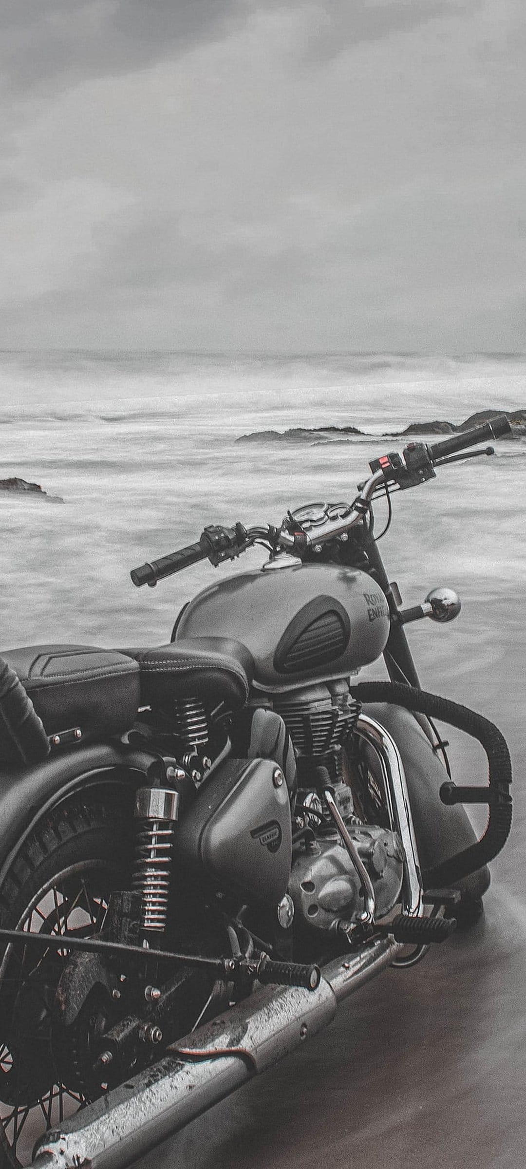 Vintage Royal Enfield motorcycle parked on wet sand with misty ocean waves crashing in a monochrome composition.