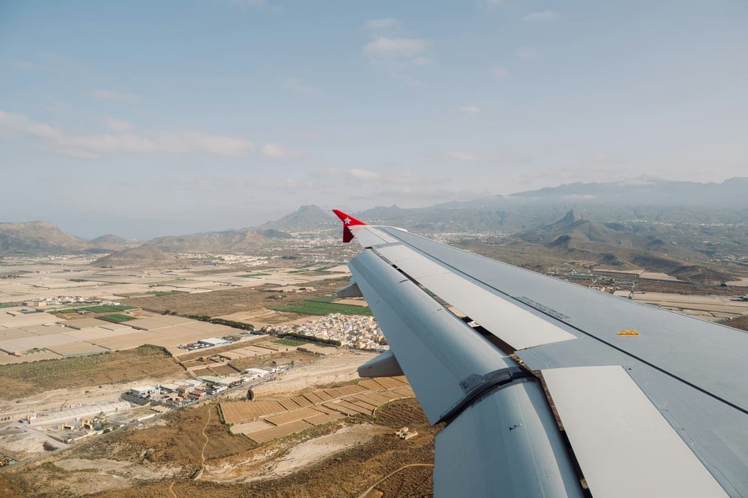 Airplane wing points toward an expansive arid landscape with geometric fields and hazy distant mountain ranges.