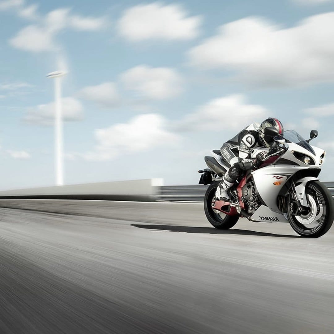 Yamaha motorcycle rider in white and red leathers leans into a racetrack curve under a blue sky with wind turbines.