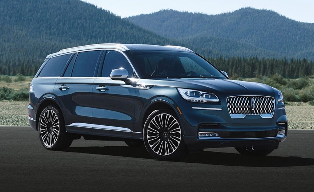 Metallic dark blue Lincoln Aviator SUV on a road with a backdrop of pine forests and blue mountains.
