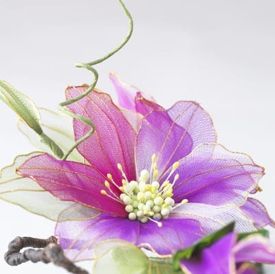 Sheer purple fabric flower petals with gold trim, yellow stamen, and green leaves on a clean white backdrop.