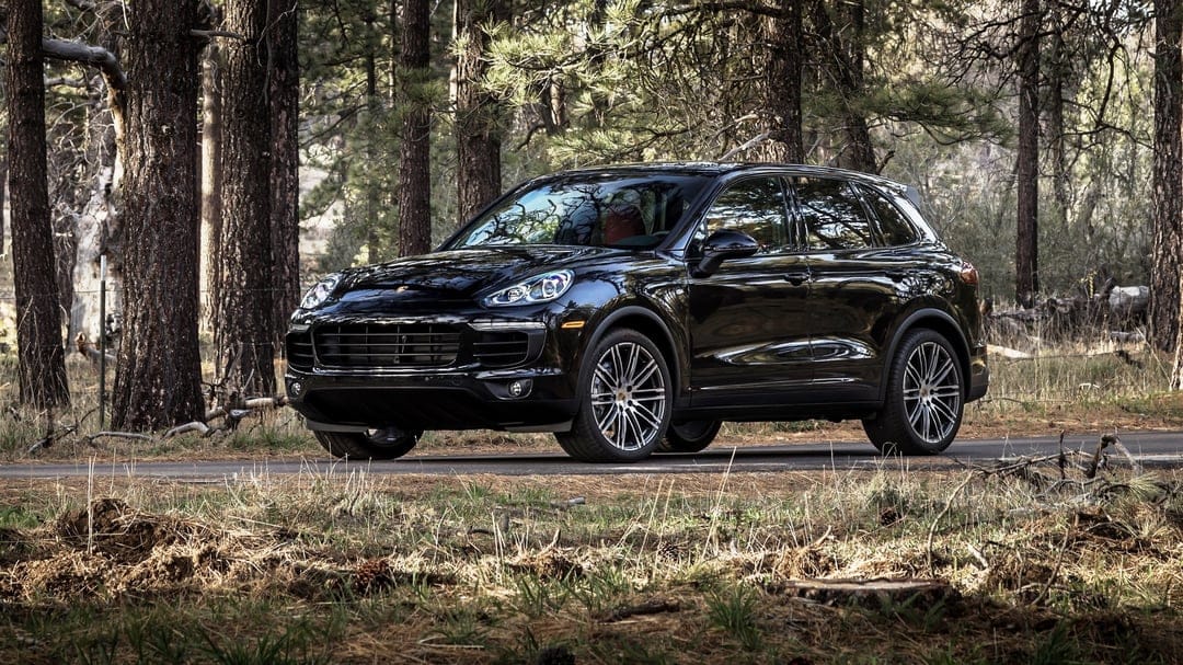 Black Porsche Cayenne SUV parked on a forest road with tall pine trees and dappled sunlight reflections.