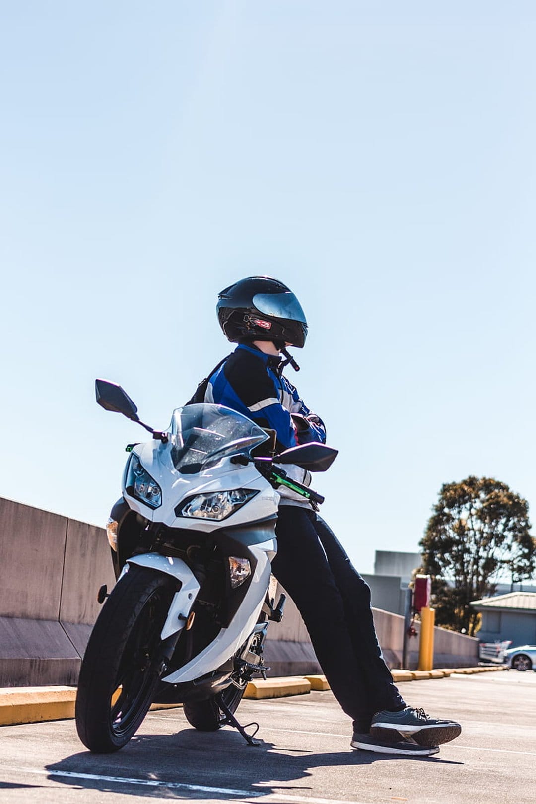 Motorcyclist in a black helmet and blue jacket leans on a white sportbike under a bright blue summer sky.