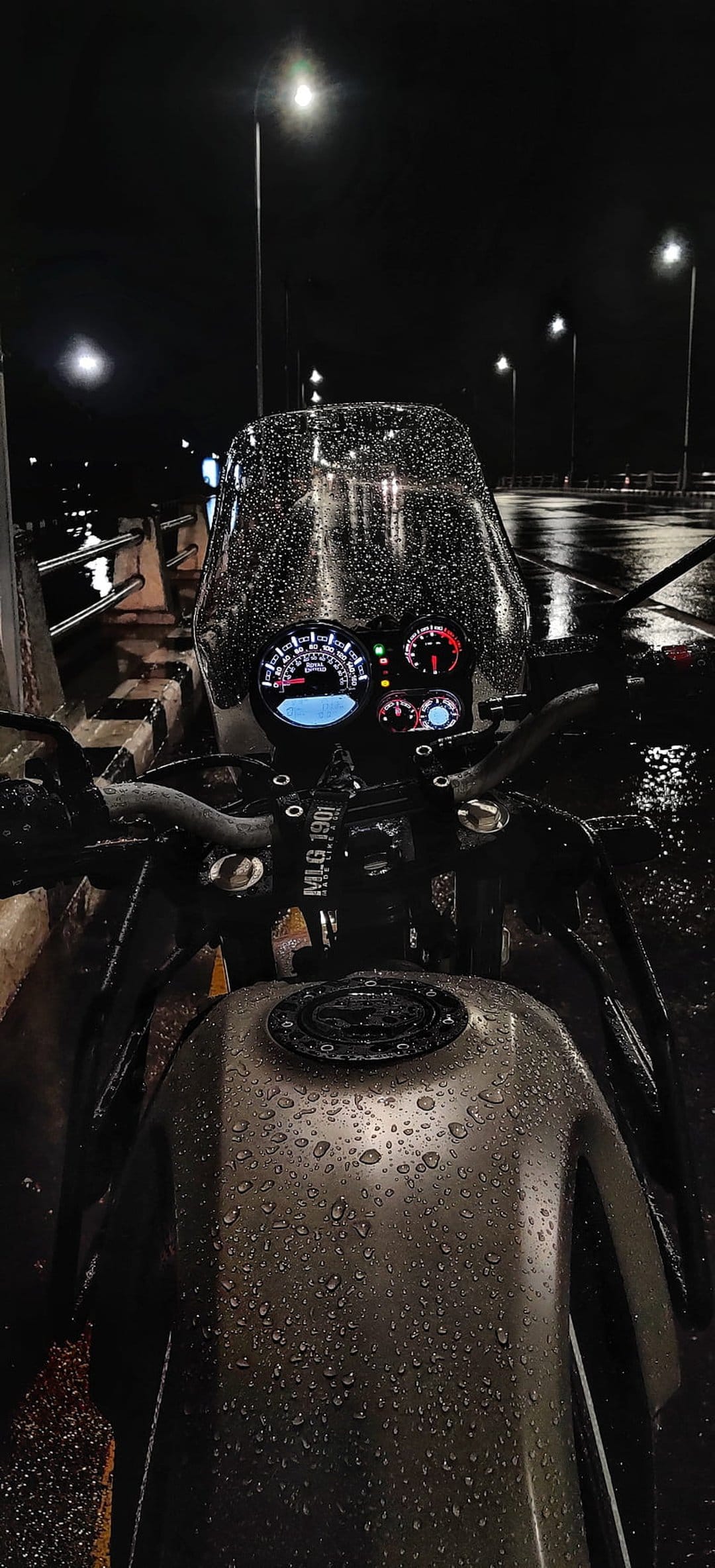 Motorcycle dashboard and fuel tank covered in rain droplets with glowing gauges and bokeh streetlights at night.