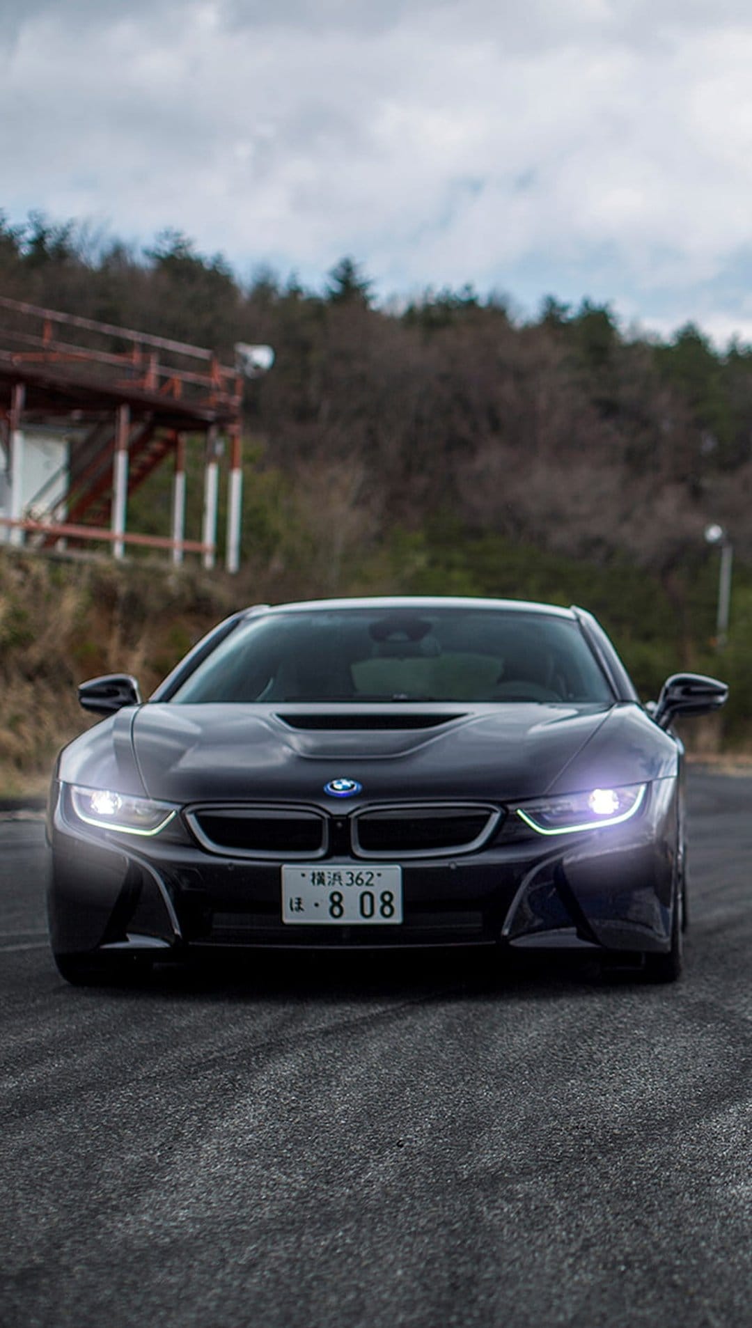 Dark BMW i8 sports car driving head-on through a curve with glowing headlights under a gloomy overcast sky.