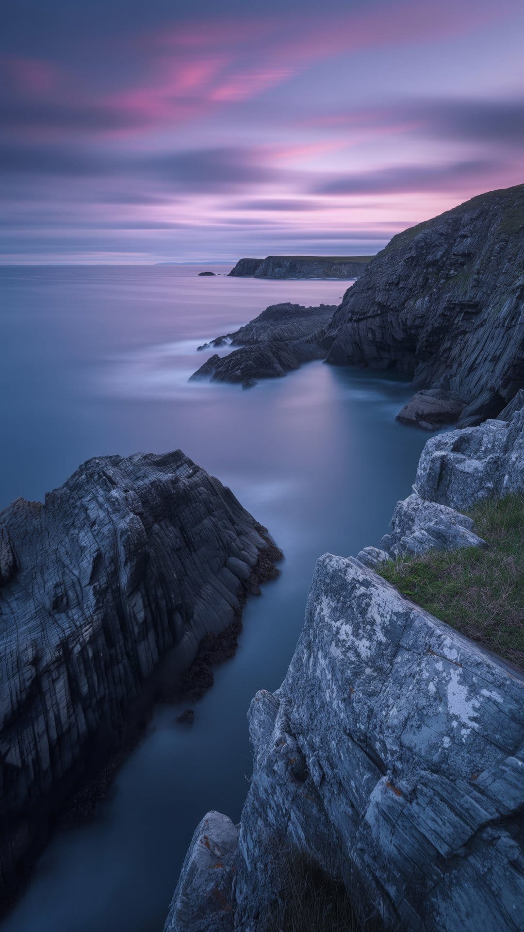 Pink and purple clouds stretch over a misty coastline where blurred waves wash against dark jagged shore rocks.