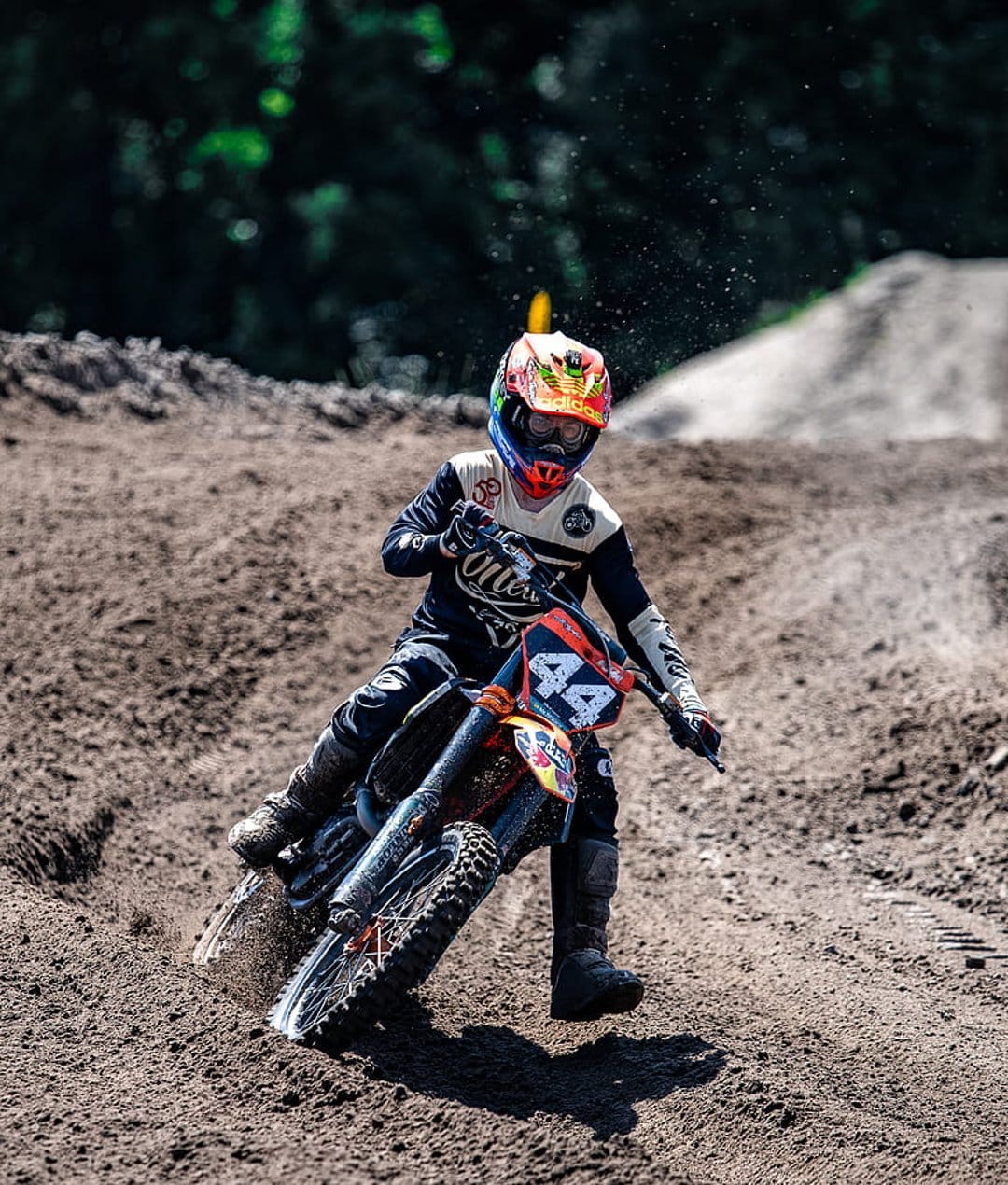 Motocross rider on an orange motorcycle leans into a dirt track turn with trees blurred in the background.