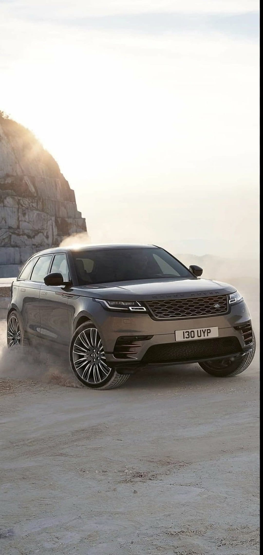 Range Rover Velar SUV driving on a dirt track, kicking up a dust cloud against a rocky cliff in hazy sunlight.