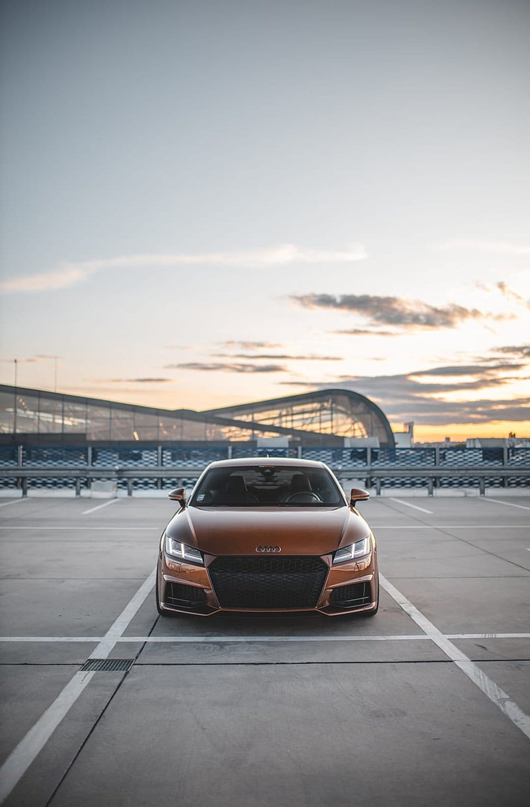 Orange Audi TT parked on a rooftop with headlights on against a sunset sky and curved modern building.