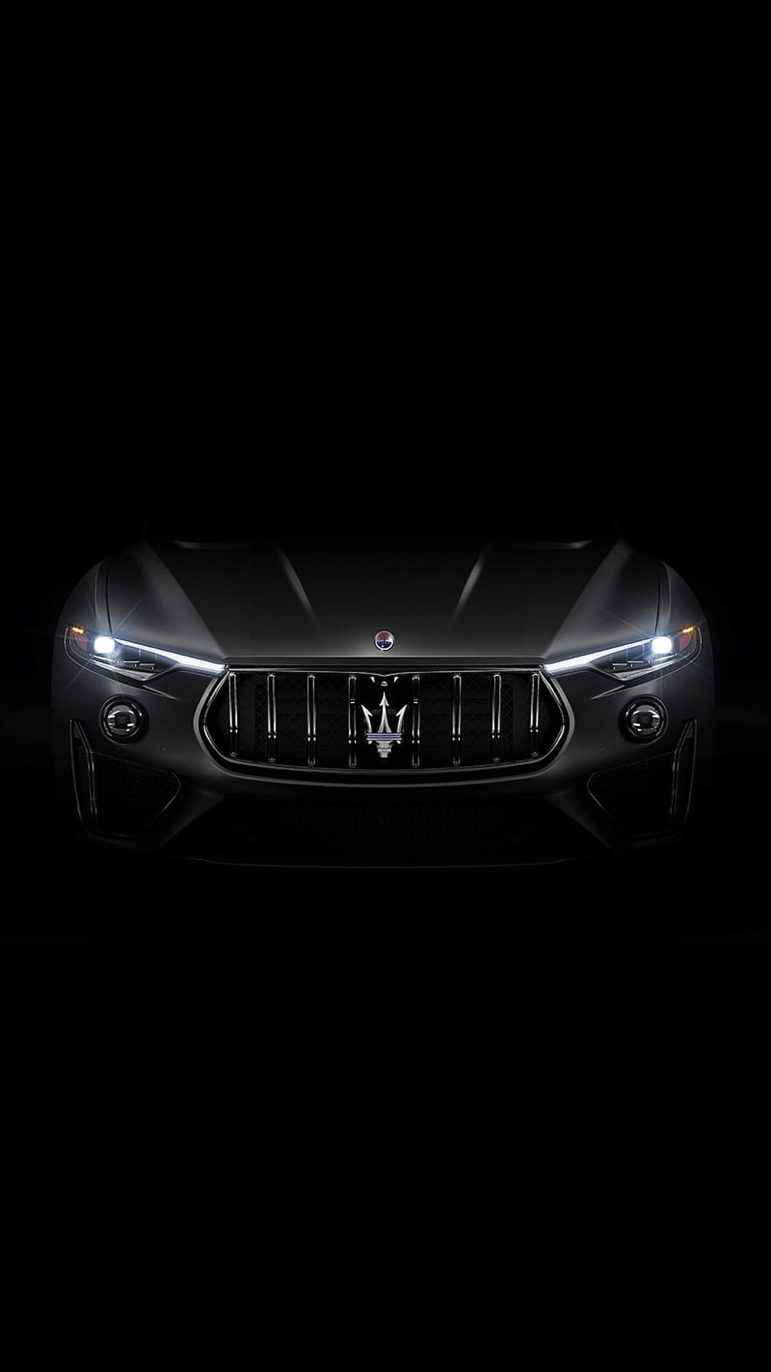 Maserati SUV front grille with chrome trident emblem and glowing headlights against a pitch black background.