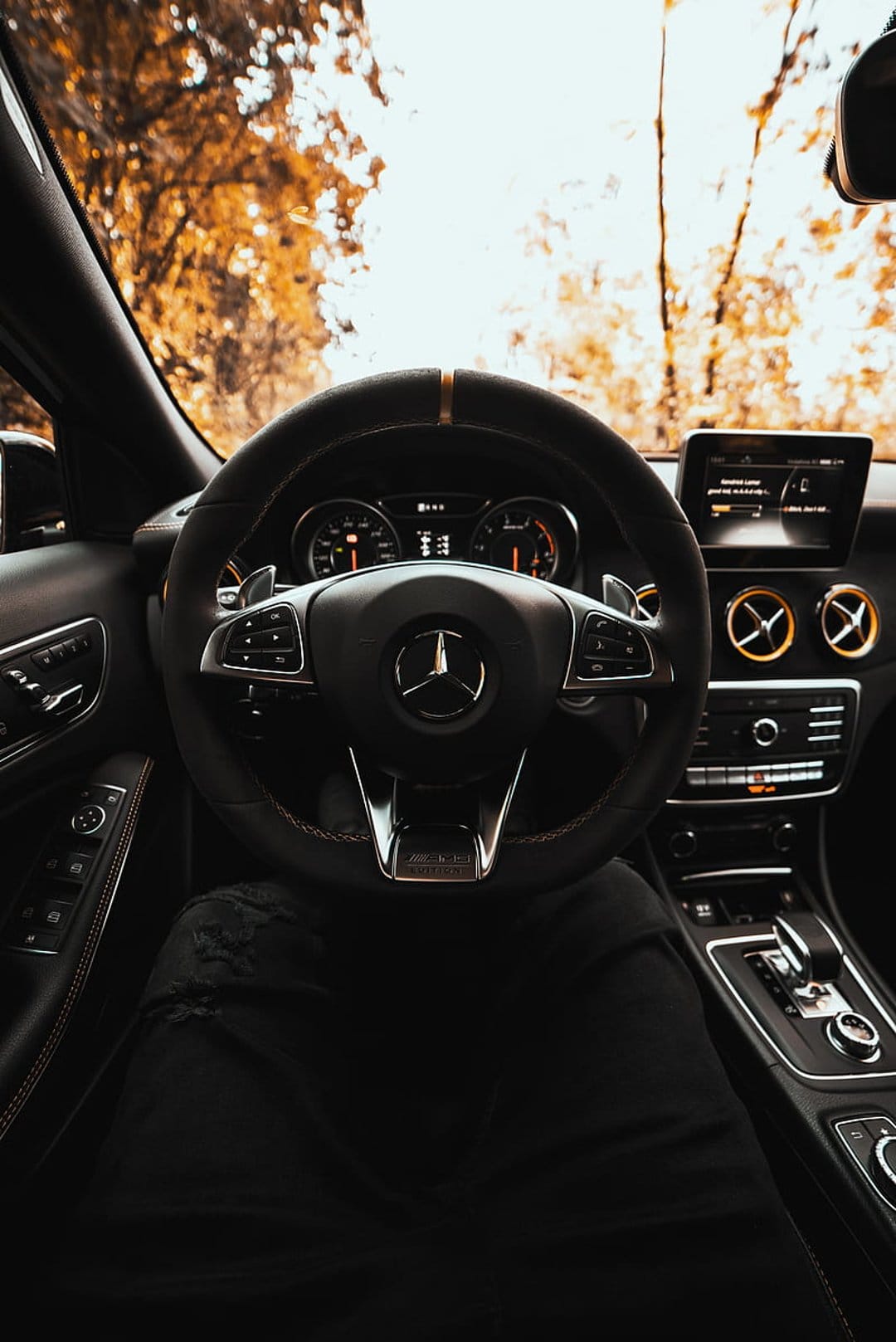 Mercedes-AMG steering wheel and digital dashboard overlook a golden autumn forest road from the driver's seat.