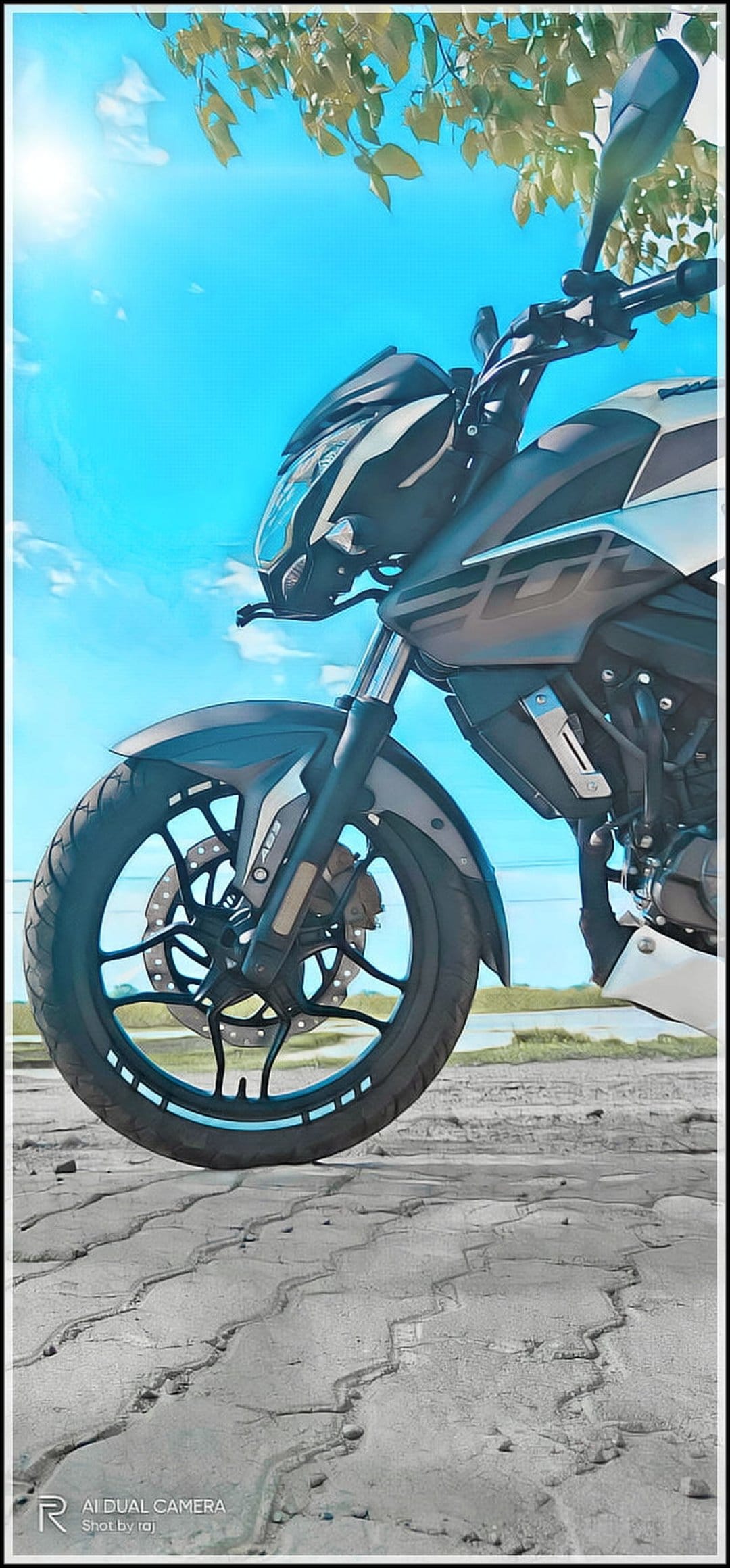 Front wheel and handlebars of a black sport motorcycle parked on pavement with green trees and a blue sky.