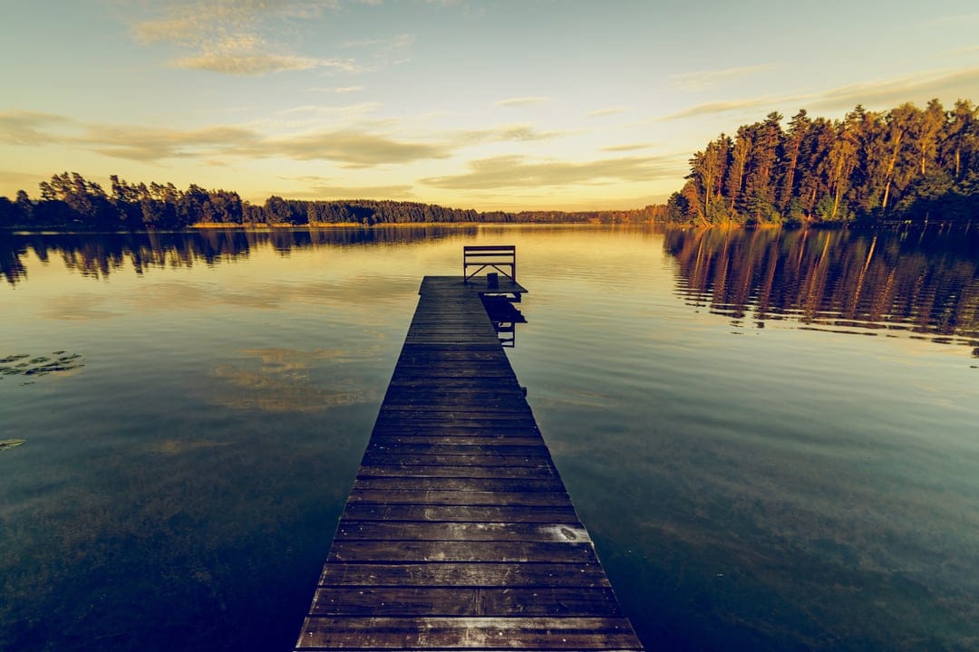 Wooden dock with a bench extends into a glassy lake reflecting an orange sunset sky and a distant forest line.