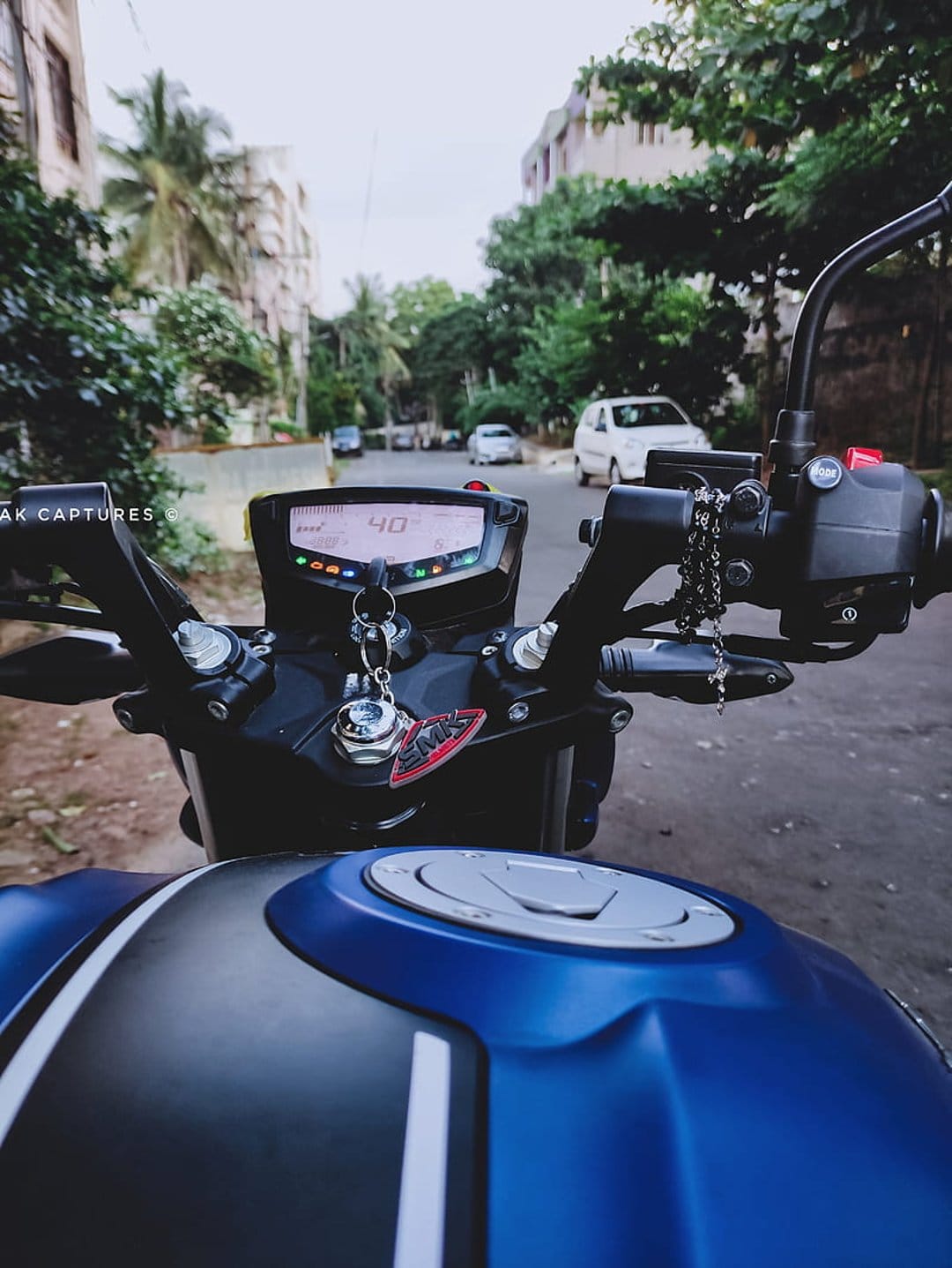 Matte blue motorcycle fuel tank and digital speedometer display on a sunlit urban street with blurred trees.