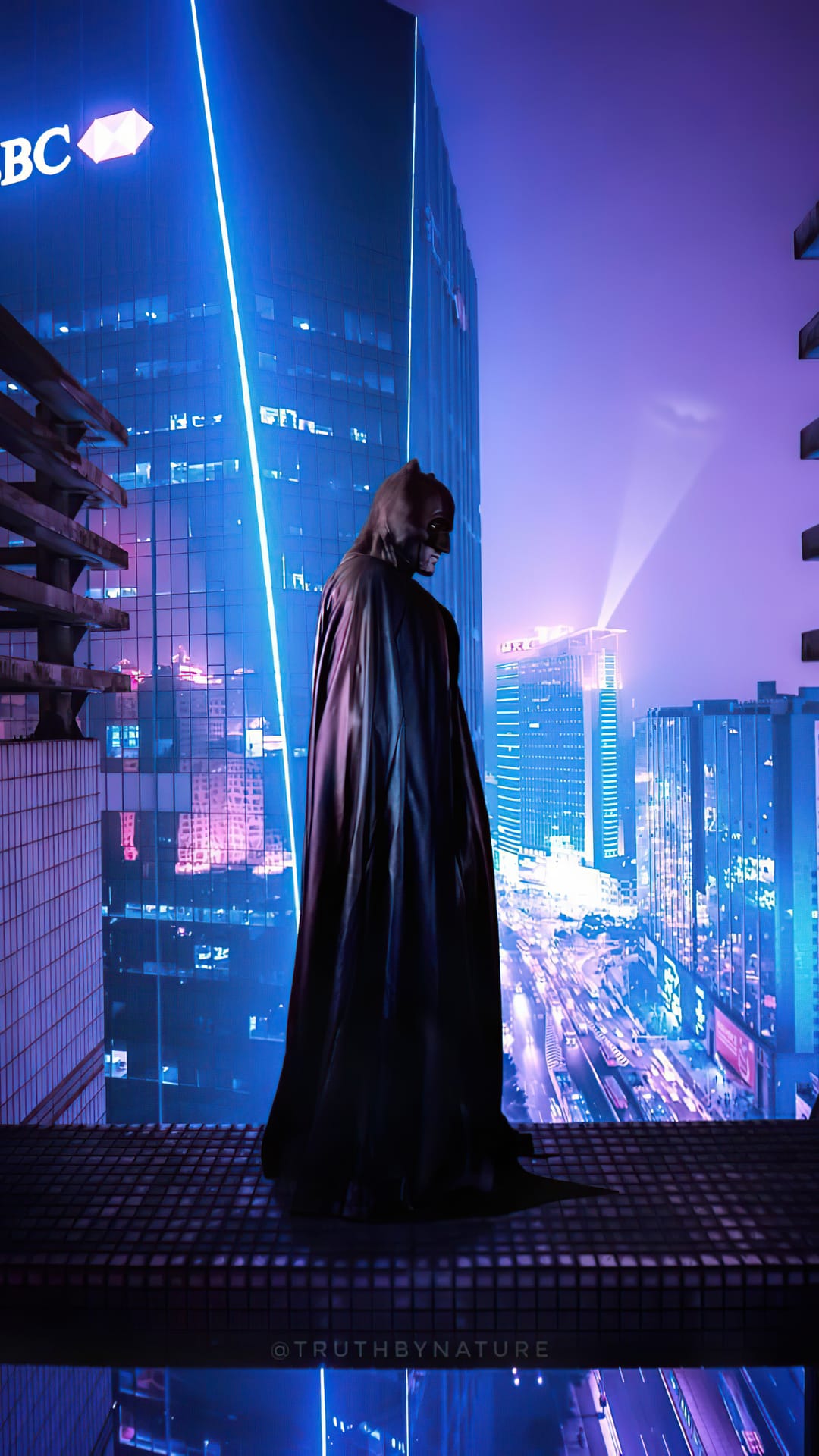Batman stands on a rooftop overlooking a glowing neon cityscape with blue and purple skyscraper lights at night.