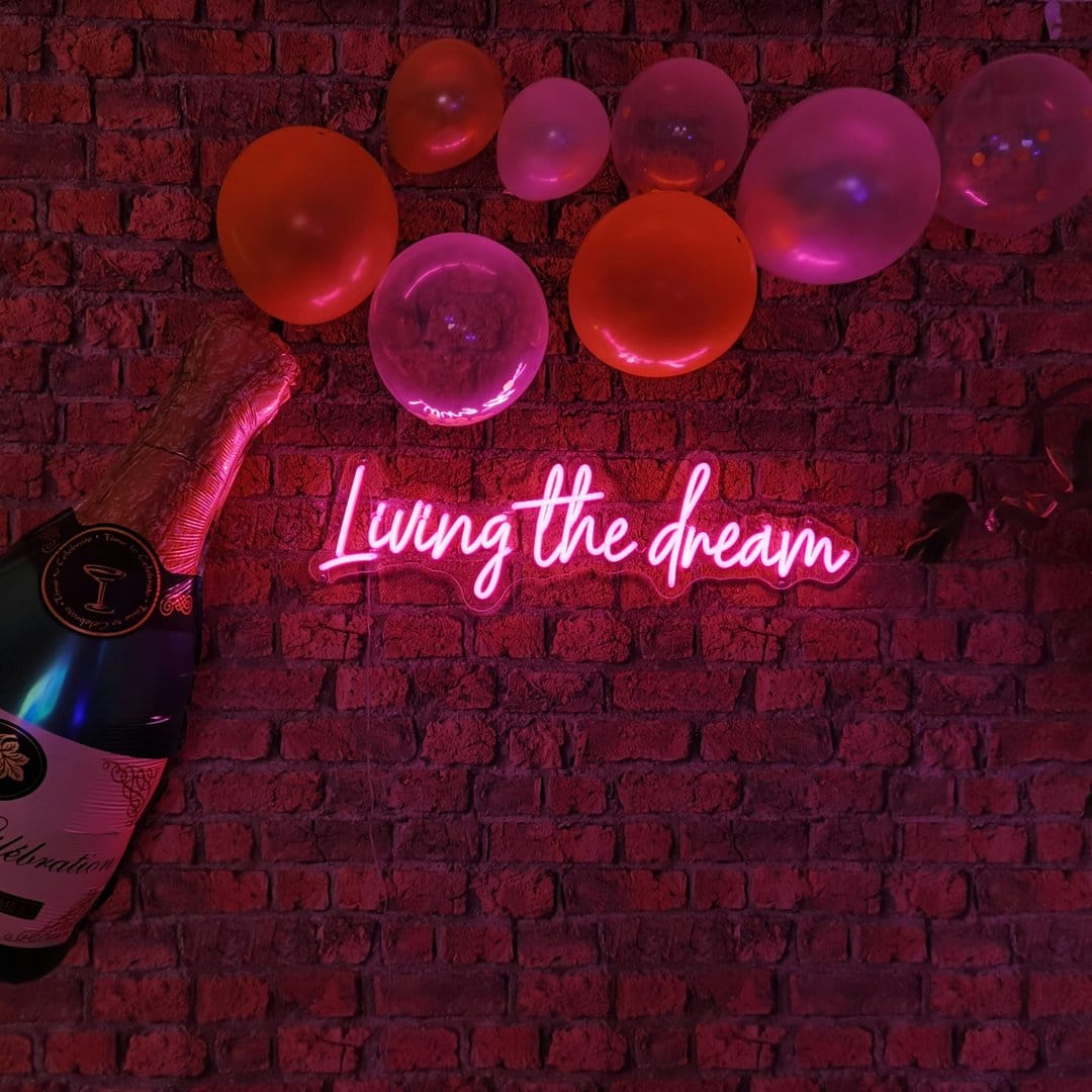 Pink neon sign reading Living the Dream glows on a brick wall next to a champagne bottle and festive balloons.