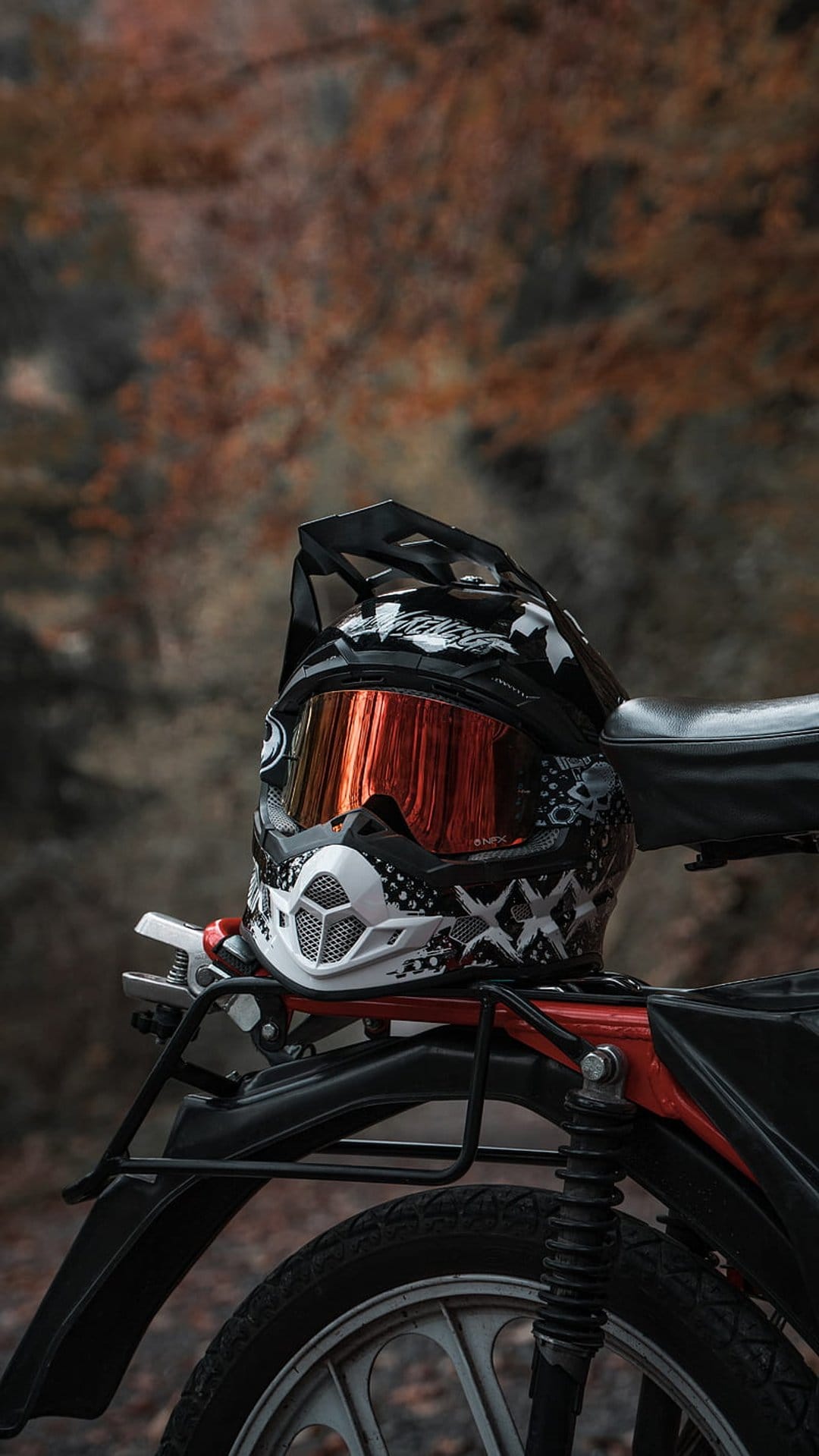 Black dirt bike helmet with mirrored goggles rests on a red motorcycle frame with orange autumn foliage behind.