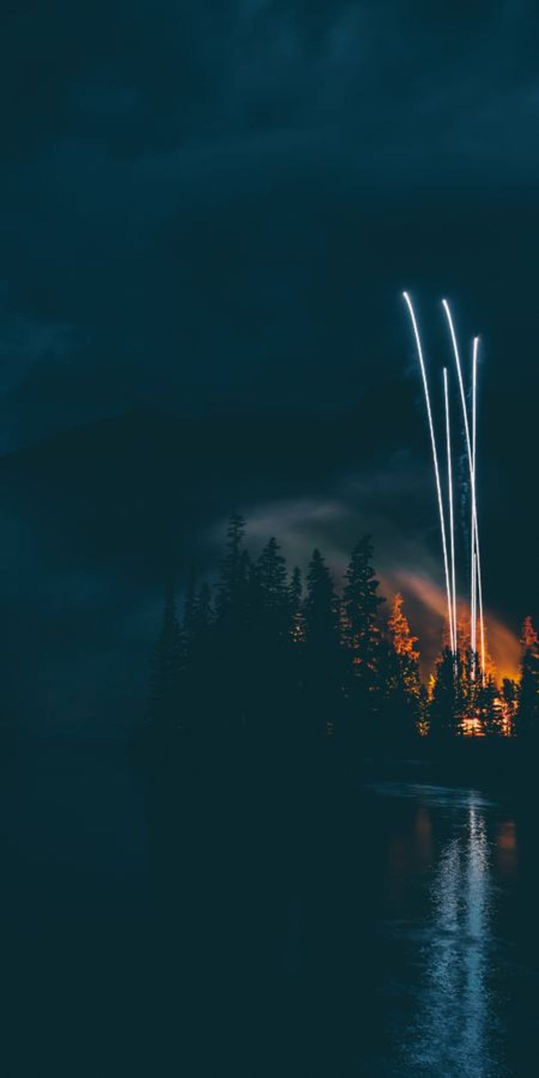 White light beams ascend from a glowing orange forest into a cloudy night sky reflected on calm lake waters.