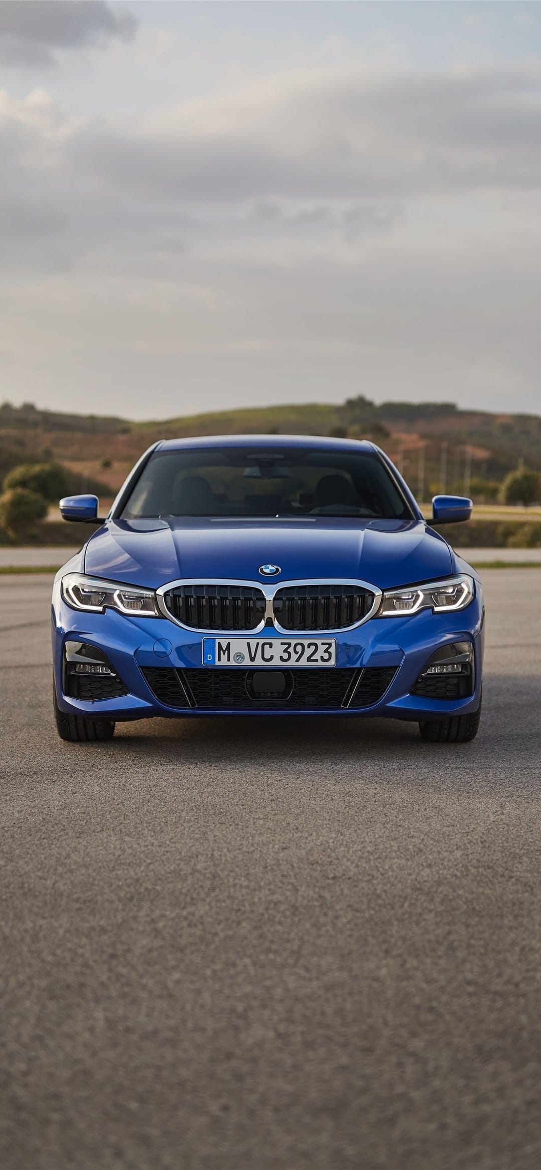 Frontal view of a metallic blue BMW 3 Series with LED headlights on asphalt under a cloudy sky.