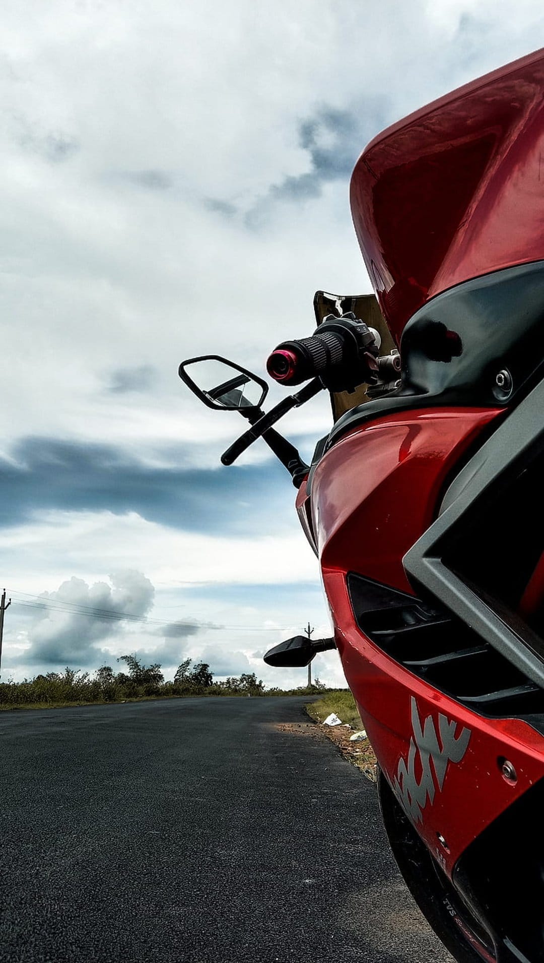Red sportbike parked on an asphalt road under a dramatic cloudy sky with green shrubs in the background.