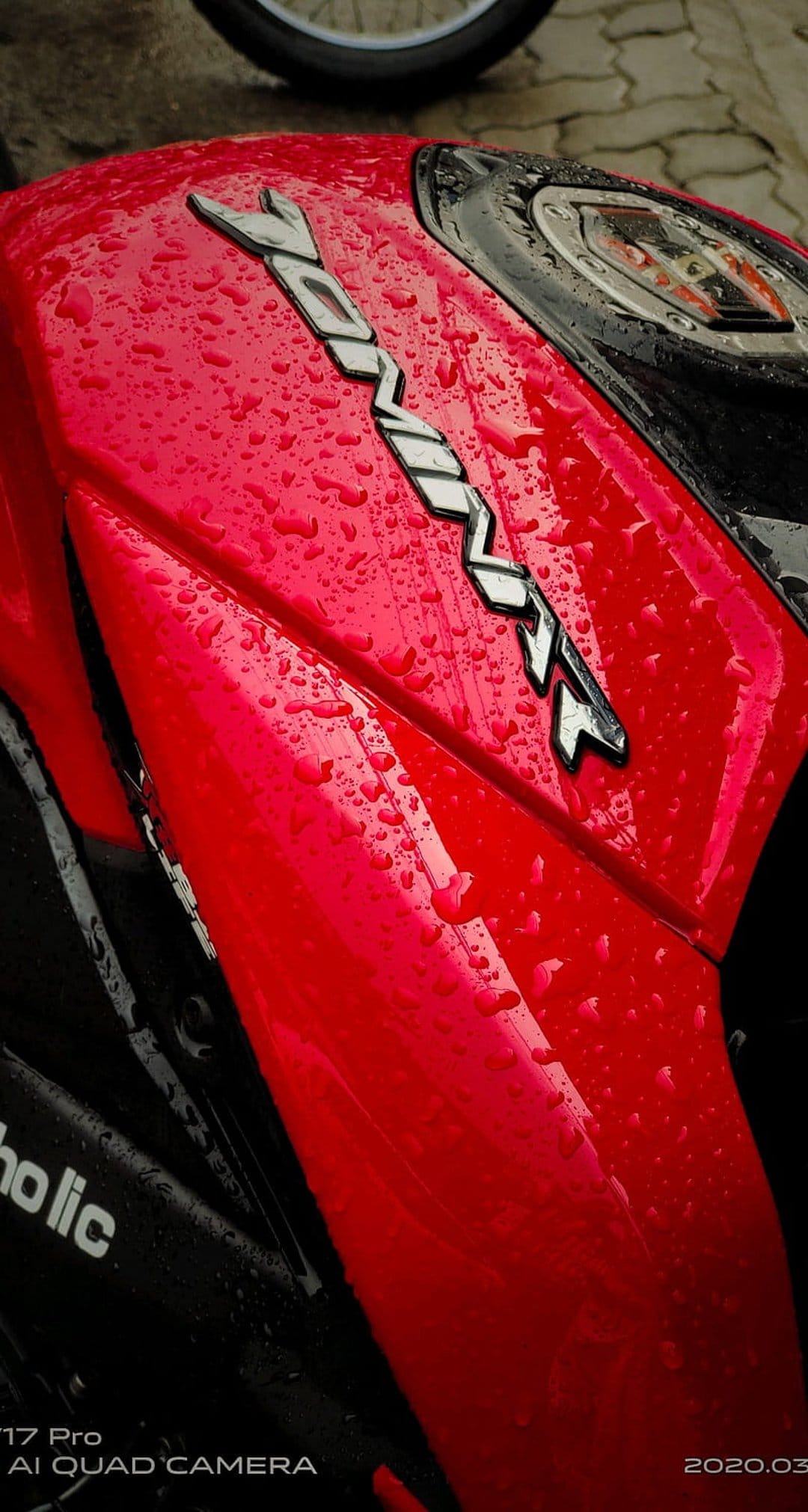 Red motorcycle fuel tank covered in water droplets with a silver chrome emblem reflecting its surroundings.