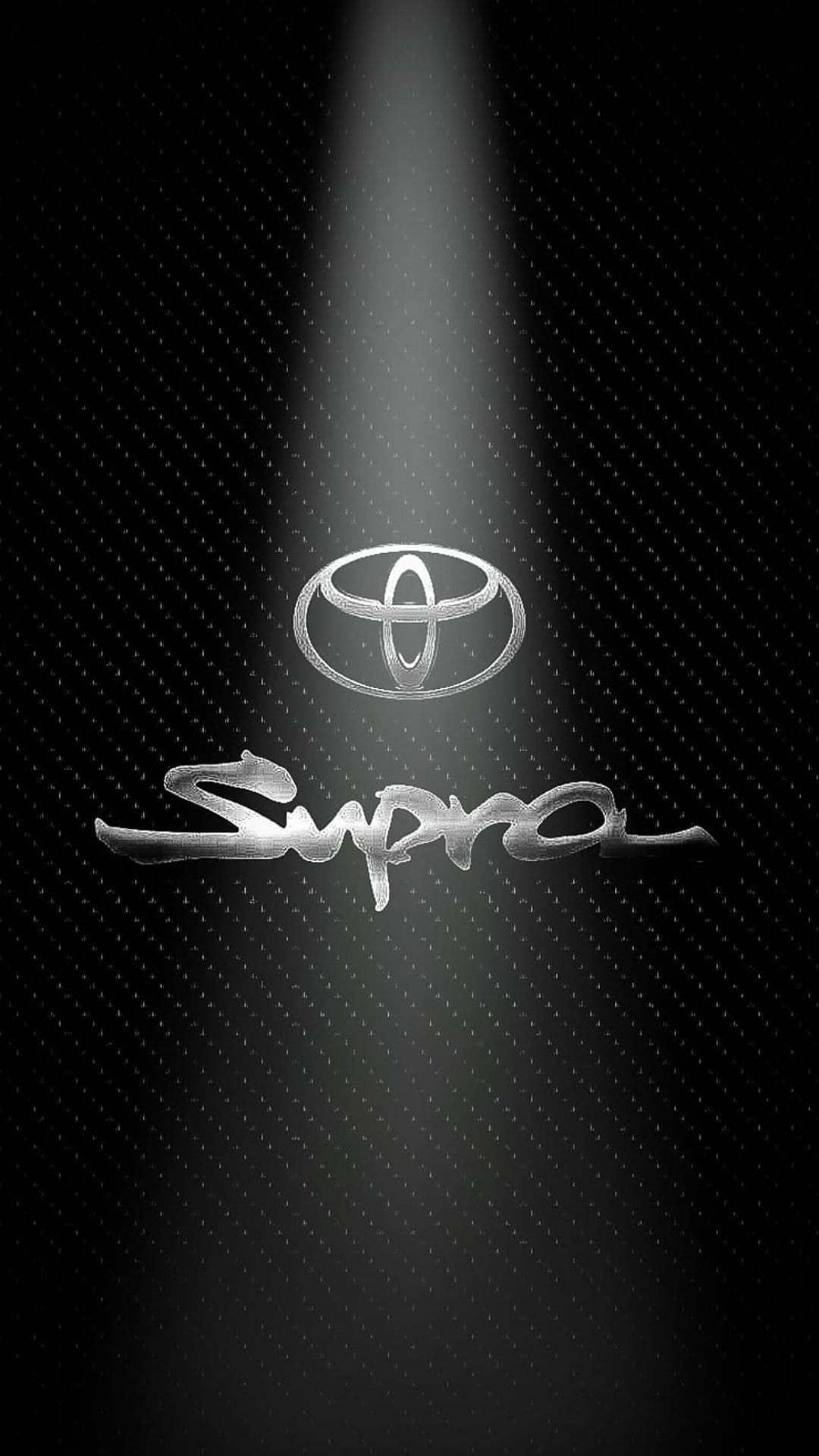 Toyota Supra chrome emblem and lettering illuminated by a spotlight on a dark textured carbon fiber background.
