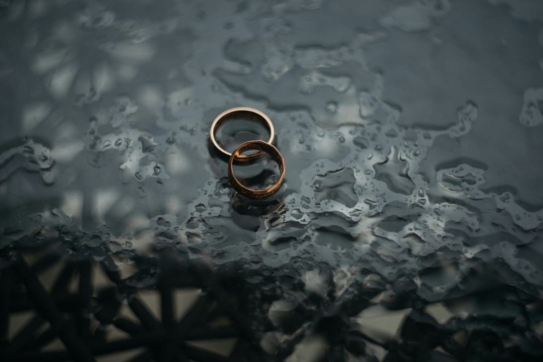 Two gold wedding rings lie on a dark wet glass surface covered in small translucent water droplets and reflections.