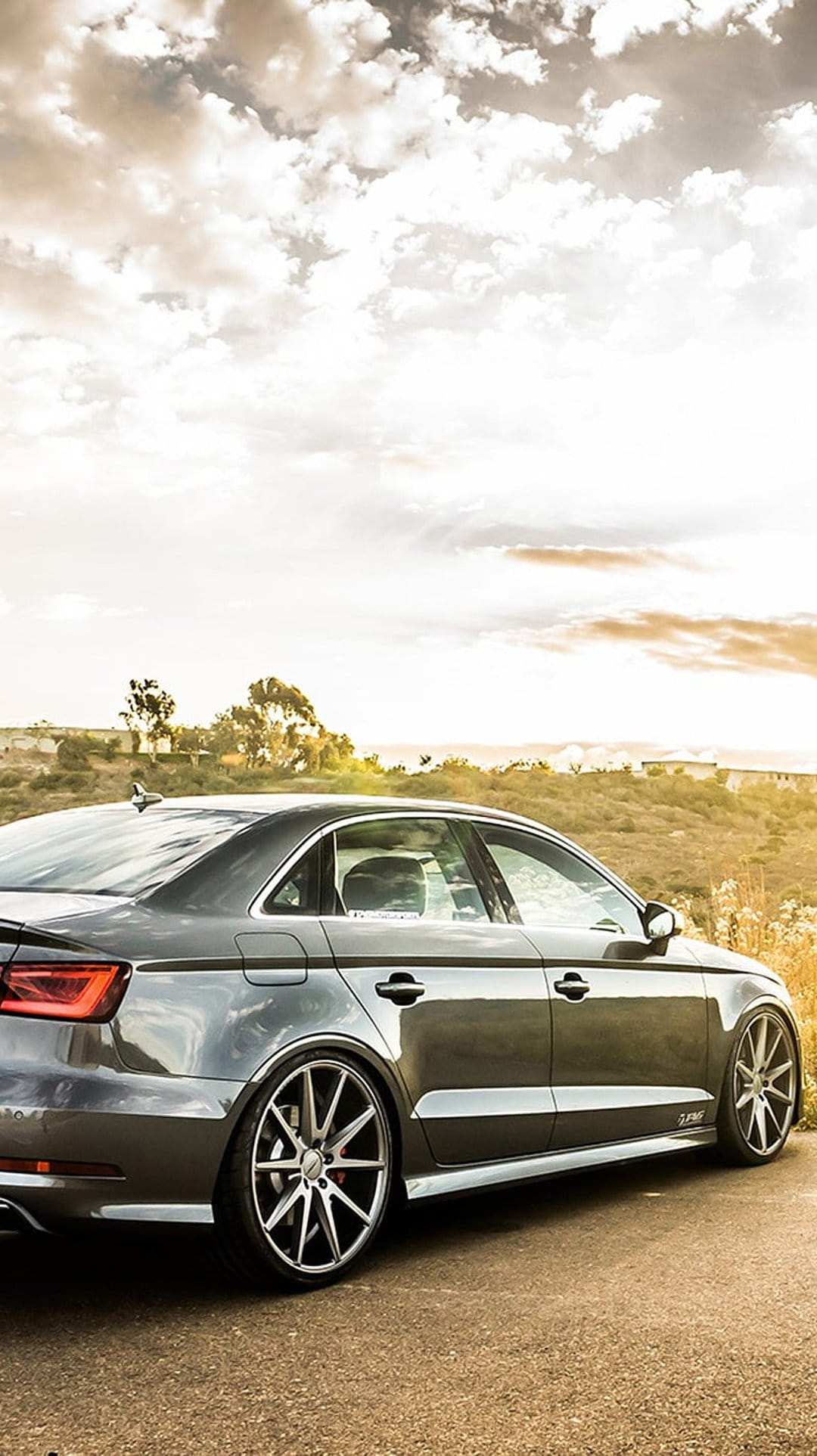 Gray sedan with multi-spoke wheels parked on a roadside during a golden sunset with rolling hills in the background.