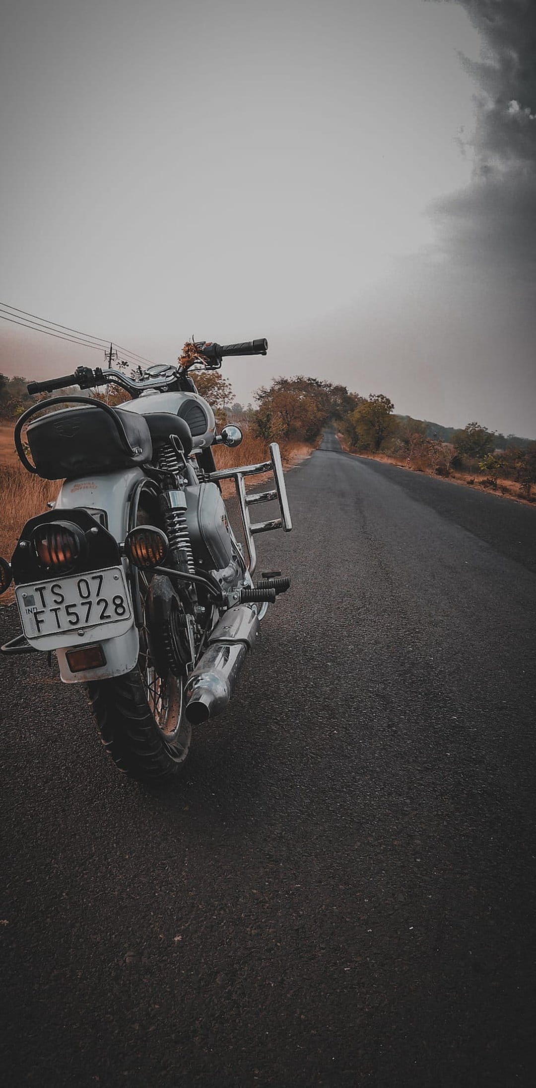 Royal Enfield motorcycle parked on an empty rural road under a dark overcast sky with dry autumn vegetation.