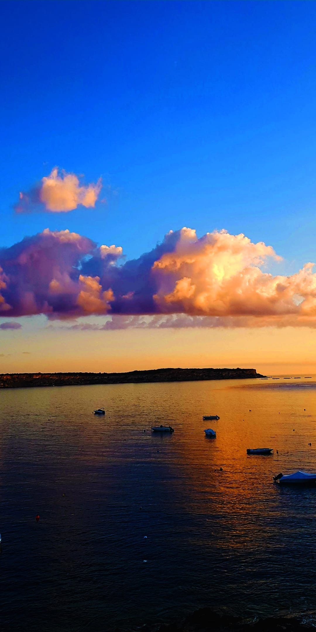 Small boats anchor in a calm bay under a dramatic orange and purple sunrise with fluffy clouds reflected in water.