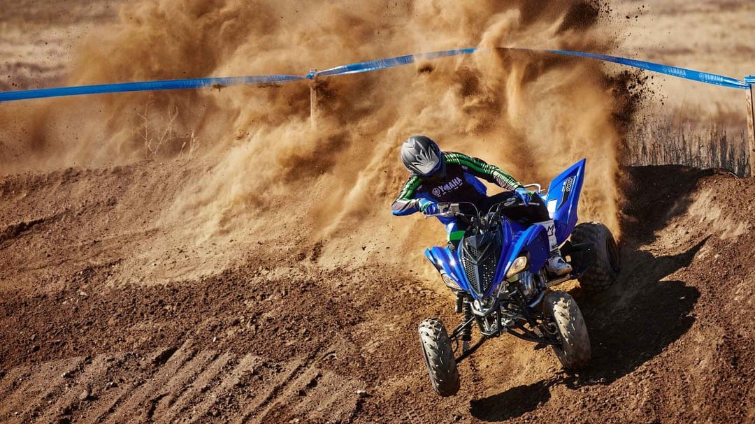 Blue Yamaha sport ATV cornering on a dirt track, kicking up a massive cloud of dust under bright sunlight.