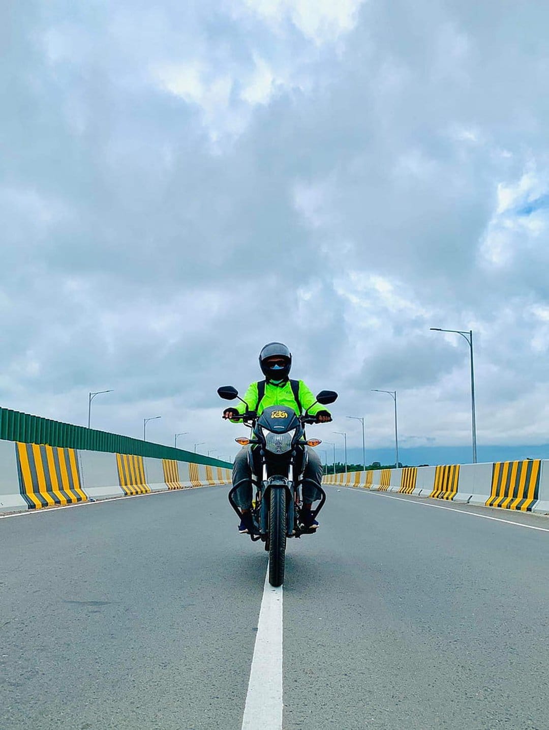 Motorcyclist in a neon green jacket riding on a highway under a dramatic cloudy sky with yellow road barriers.