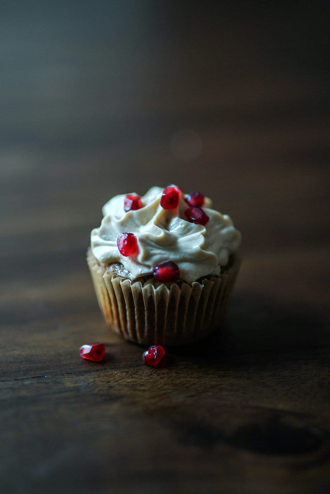 Frosted cupcake with red pomegranate seeds on a brown paper liner atop a dark rustic wooden surface.