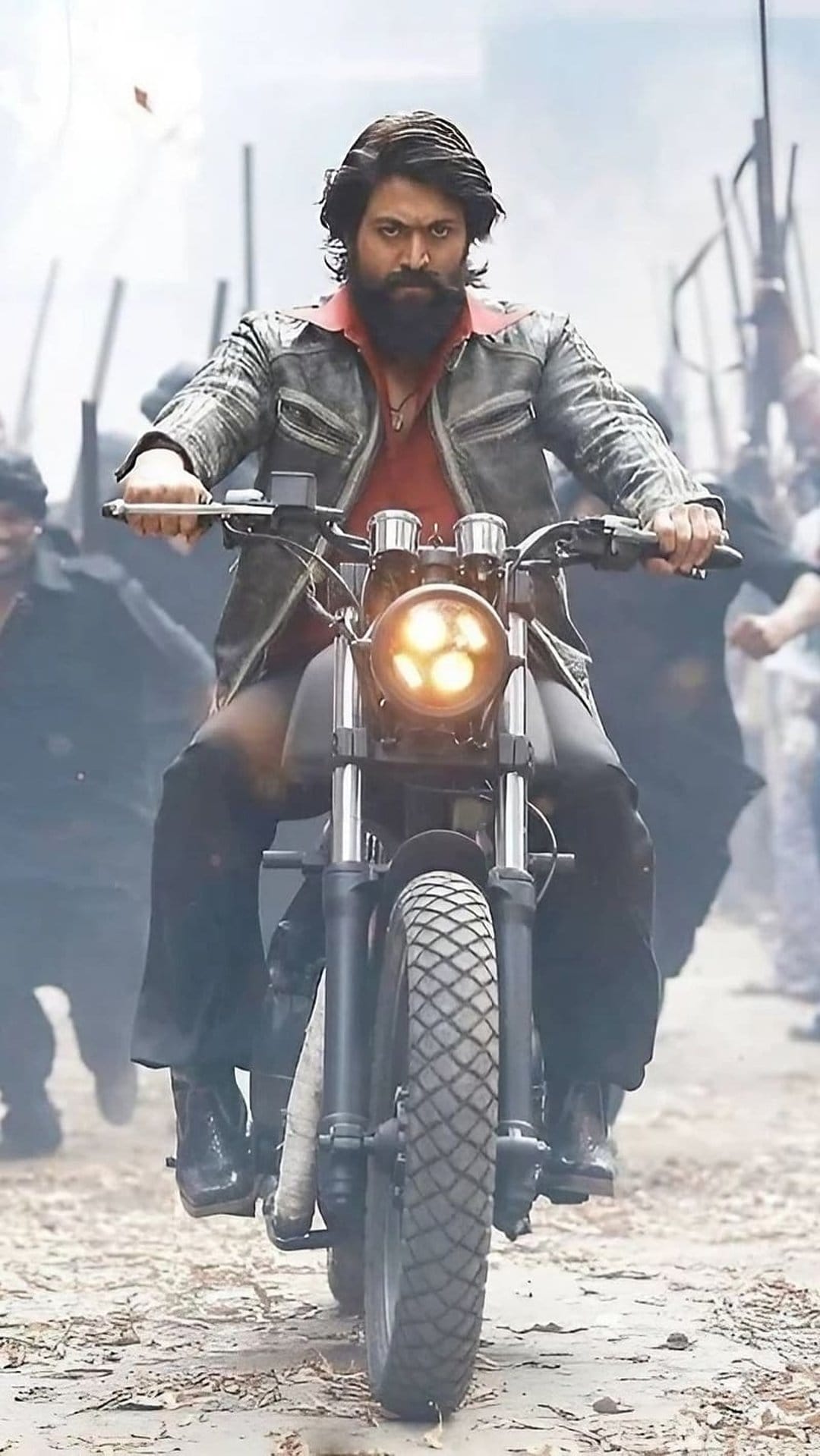 Yash as Rocky Bhai rides a motorcycle wearing a leather jacket and red shirt with a blurred crowd in the background.